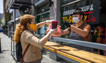 A Mexican restaurant adapts to the Covid-19 lockdown. The owner hands an order to a customer outside the restaurant; they are both wearing gloves and masks.